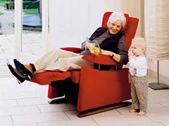 Ältere Frau sitzt gemütlich vor dem Fenster im Wohnzimmer im Pflegesessel mit hochgestellter Fußstütze und rotem Stoffbezug und spricht mit ihrem kleinen Enkel.