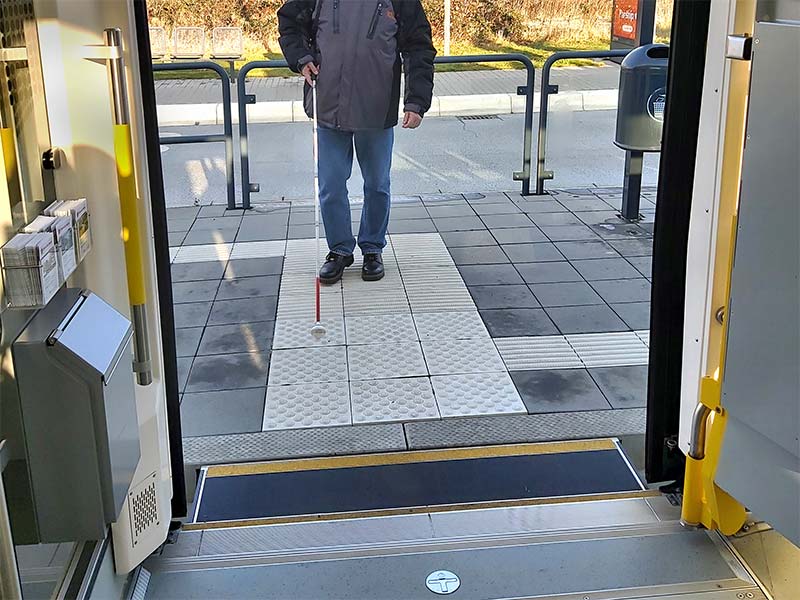 Mensch mit Langstock geht auf dem Leitsystem zur Tür der Straßenbahn