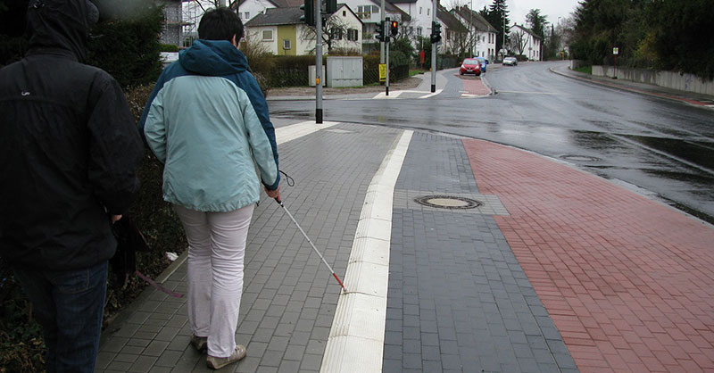 Blinde Frau mit Taststock auf dem Geeg entlng der Radwegeplatte.