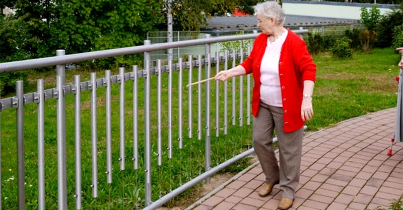 Seniorin läuft mit einem Stöckchen an einem Klangzaun entlang. 