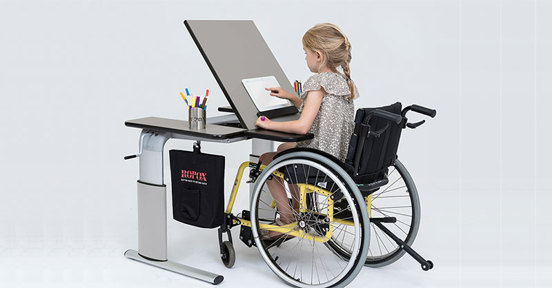 Kind im Rolli sitzt am Tisch mit geneigter Tischplatte und liest
