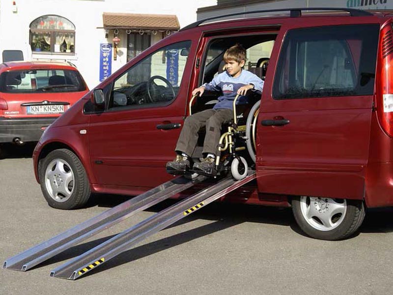 Junge fährt mit seinem Rollstuhl aus der Seitentür eines Autos die Rampen hinunter