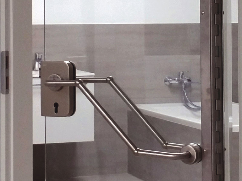 Türdetail einer Ganzglastür mit metallfarbener barrierefreier Drückergarnitur,  mit Durchblick zum Badezimmer