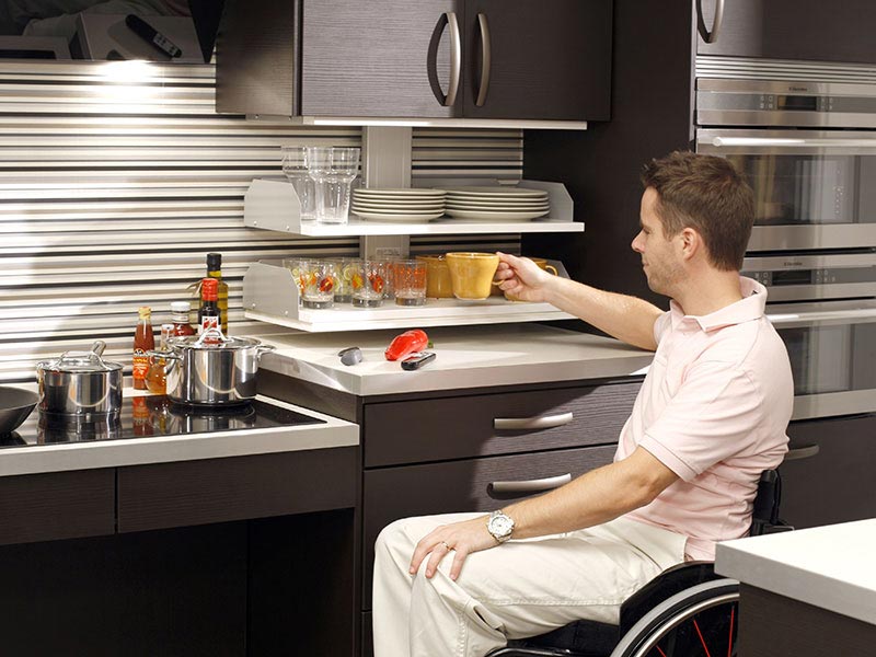 Rollstuhlfahrer entnimmt Geschirr aus abgesenktem Küchenoberschrank