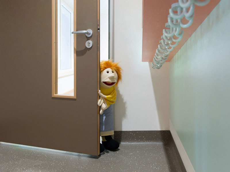 Durch die  geöffnete Eingangstür einer Kita schaut eine Kinderpuppe im Stil der Sesamstraße