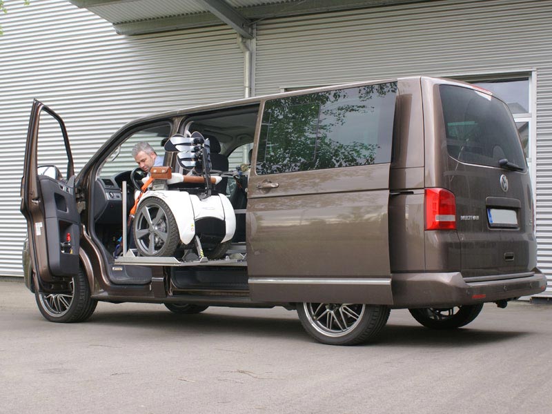 Elektrorollstuhl steht auf Hebeplattform Scooterboy, wird durch seitliche Schiebetür eines VW-Bus befördert