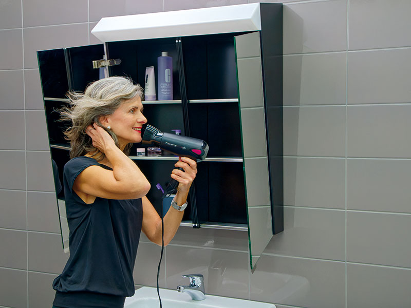 Frau fönt sich die Haare und betrachtet sich in den Innenspiegeln des Badezimmerschrankes