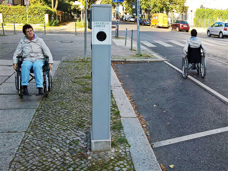 Rollstuhlfhrerin auf dem Weg vom parkenden Fahrzeug über abgesenkte Bordkante zur Ladesäule.
