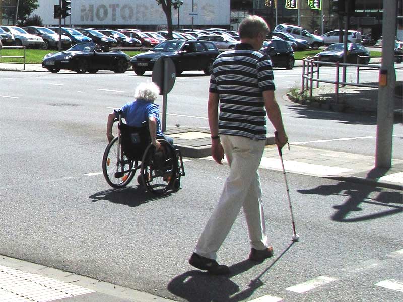 Eine Rollstuhlfahrerin und ein Blinder überqueren eine getrennte barrierefreie Querungsstelle.