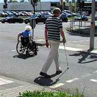 Eine Rollstuhlfahrerin und ein Blinder überqueren eine getrennte barrierefreie Querungsstelle.