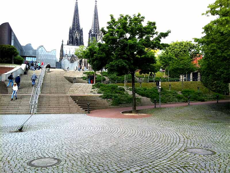 Treppe zur Domplatte in Köln. Die Führung zum Handlauf in der Treppenmitte erfolgt über ein Metallelement im Boden. Für Menschen, die auf Rollstuhl oder Rollator angewiesen sind, dient der Weg rechts als Verbindung.